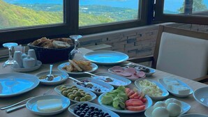 Ένα τραπέζι στρωμένο με διάφορα πιάτα, με θέα σε μια λίμνη και βουνά, που διακρίνονται μέσα από μεγάλα παράθυρα.
