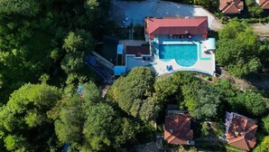 Μια αεροφωτογραφία μιας ιδιοκτησίας με μια πισίνα, ένα κτίριο με κόκκινη στέγη και την πλούσια βλάστηση που την περιβάλλει.