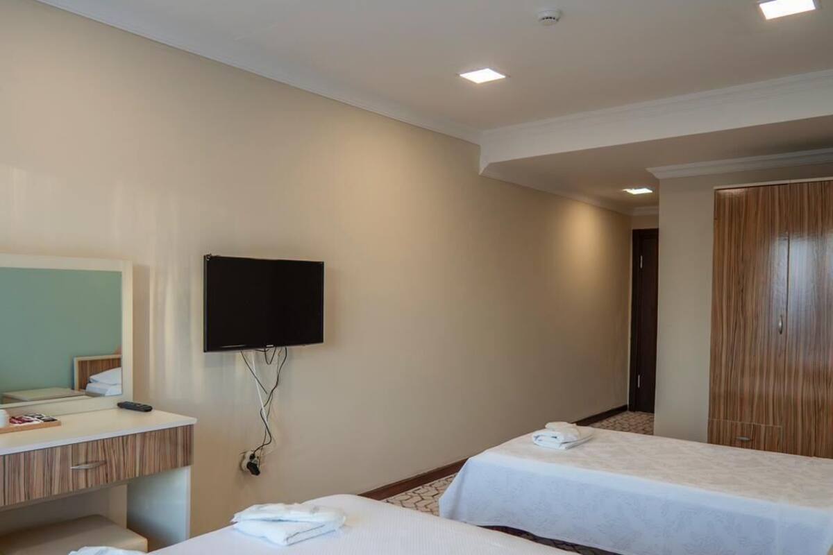 A hotel room with two beds, a TV mounted on the wall, a wooden wardrobe, and a small desk with a mirror.