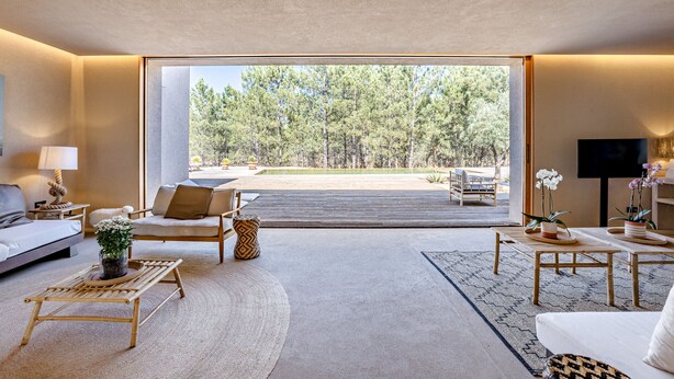 A modern living room with a large sliding glass door leading to a deck, furnished with a sofa, chairs, and a coffee table. - Melides Premium Villa by Homing (Grandola)