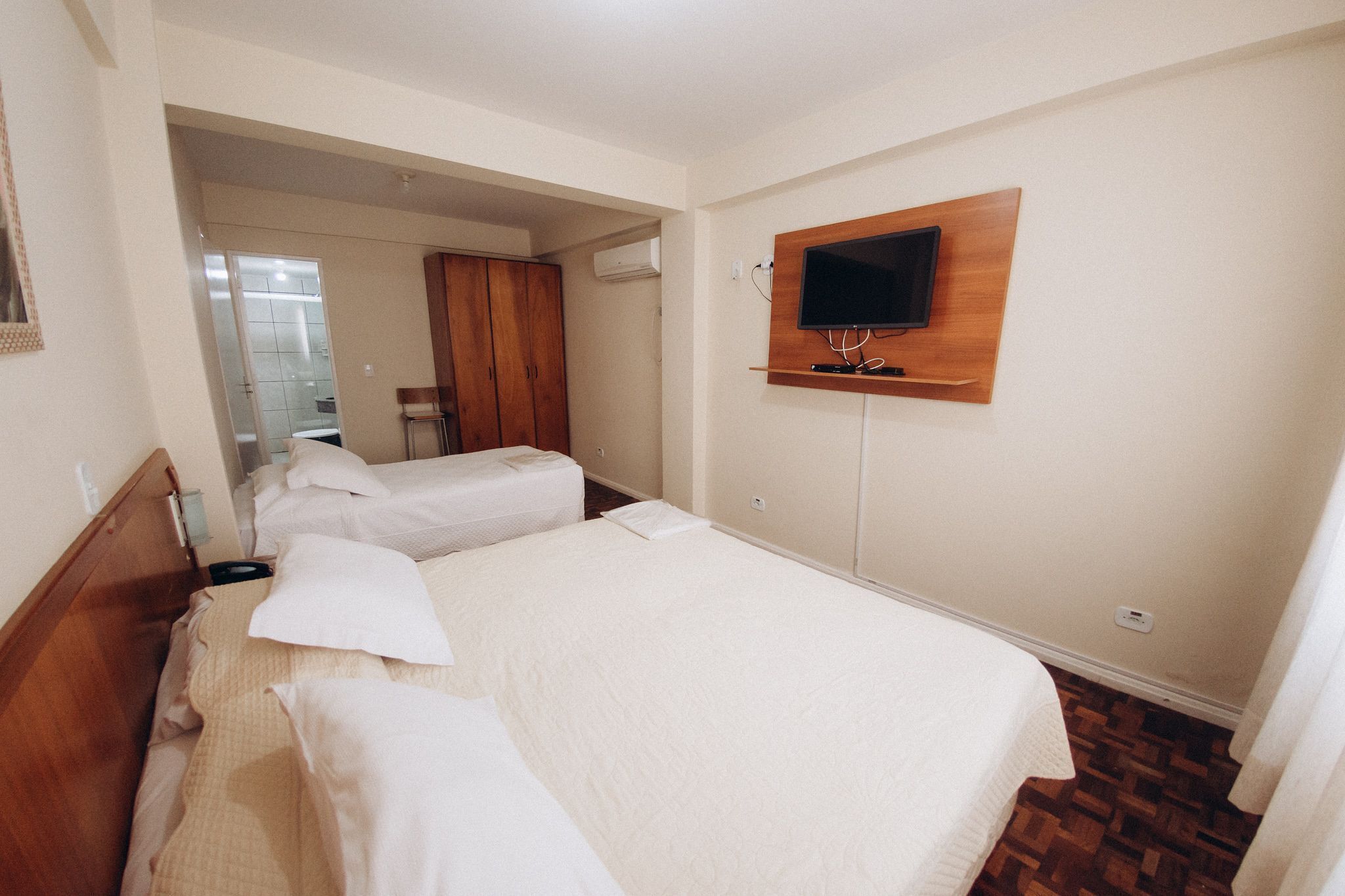 Habitación de hotel con dos camas, un armario de madera, un televisor montado en la pared y un baño visible a través de una puerta abierta.