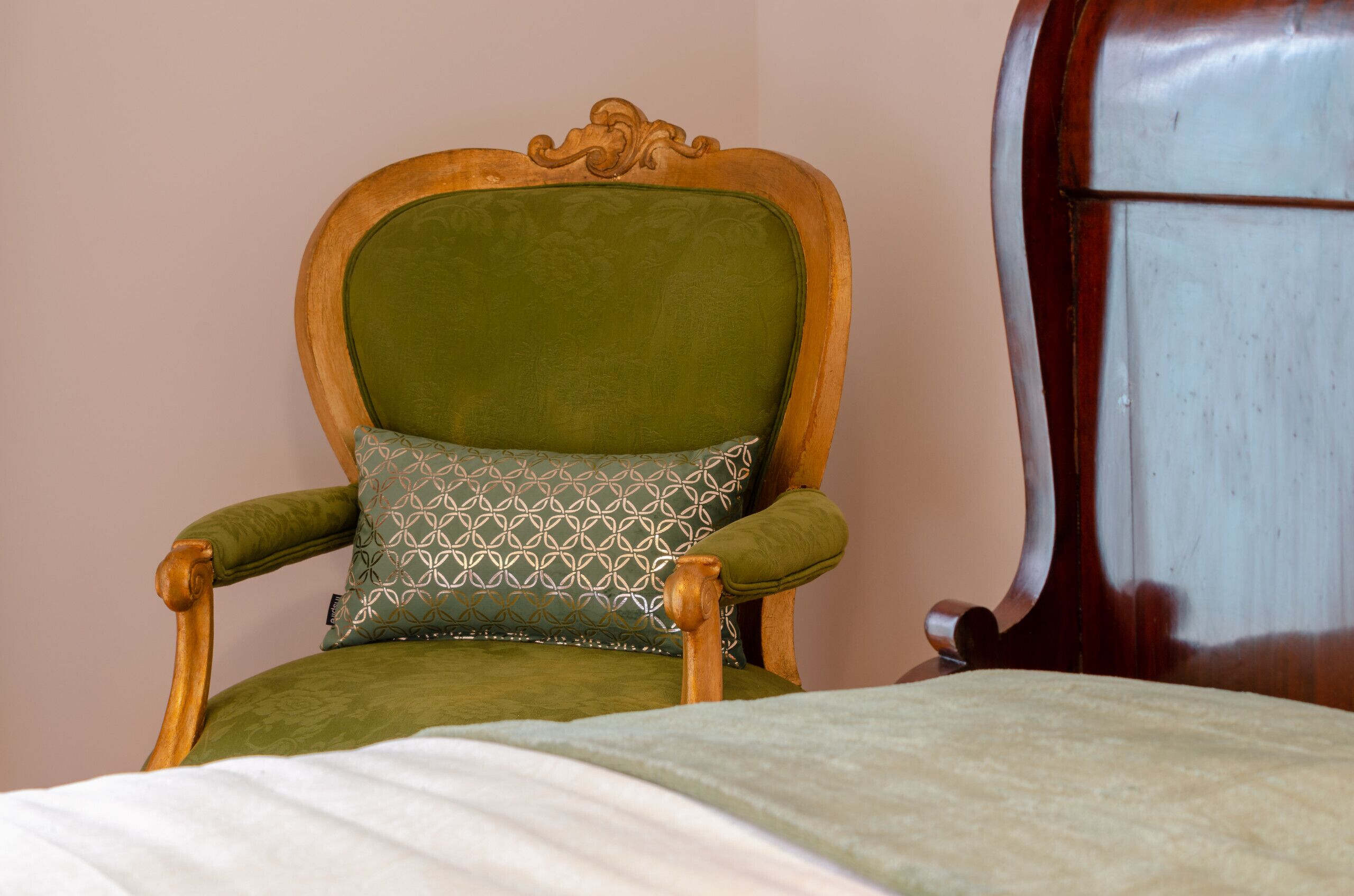 Une chaise en bois avec un dossier rembourré de tissu vert et un coussin à motifs, placée à côté d’un lit recouvert d’une couverture blanche.