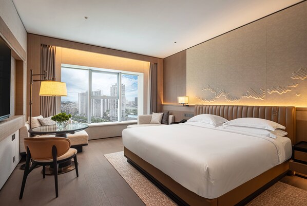 A modern hotel room with a large bed, a seating area with a table, and a view of the city through the window.
