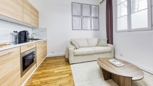 Una cocina moderna con armarios de madera, horno integrado y encimera de mármol. Se observa un sofá color beige y una pequeña mesa de madera con un libro.