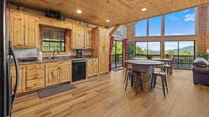 A spacious kitchen with wooden cabinets and a large island, a dining area with a round table and chairs, and a large window offering a view of the outdoors. - Five Seasons - Luxury Cabin Featuring a Pool Table, Hot Tub and Fire Pit (Cleveland)