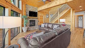 A spacious living room with a large leather sectional sofa, a stone fireplace, and a wooden staircase. - Five Seasons - Luxury Cabin Featuring a Pool Table, Hot Tub and Fire Pit (Cleveland)