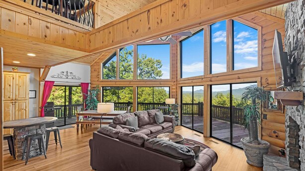 A spacious living area with a large brown leather sofa, a stone fireplace, and a view of the outdoors through large windows. - Five Seasons - Luxury Cabin Featuring a Pool Table, Hot Tub and Fire Pit (Cleveland)
