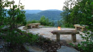 A stone fire pit with benches in a forested area with a view of distant hills.