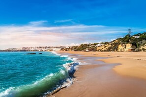 A coastal beach with clear blue waters, sandy shore, and a town in the background. - Convenient Stay, Superb Location! 2 Outdoor Pools, Free Parking, Airport Shuttle (Albufeira)