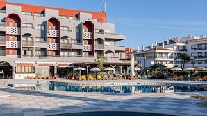 A pool area with lounge chairs and umbrellas, a building with balconies and a terrace, and a clear sky.
