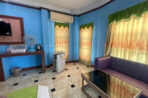 A room with a sofa, a table, a fan, a small refrigerator, and a TV on a stand.