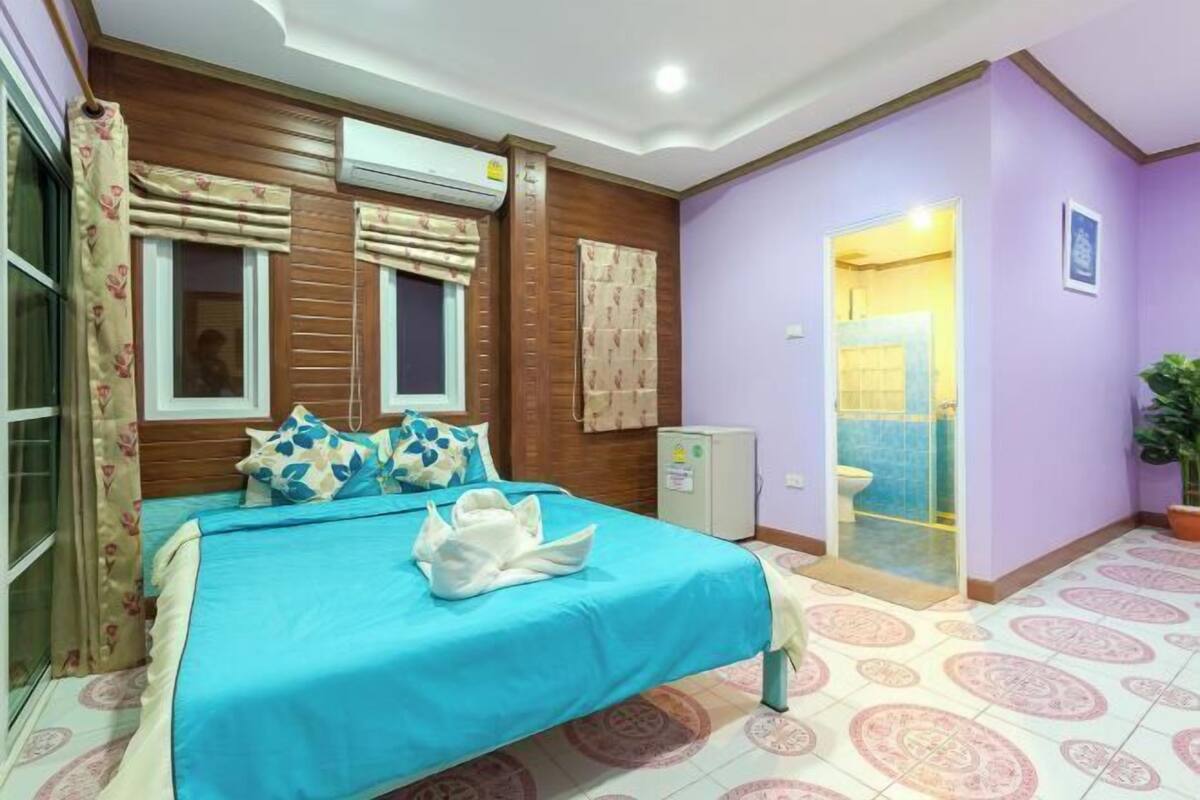 Une chambre à coucher comprenant un lit, une fenêtre avec des rideaux, un appareil de climatisation, un petit meuble et une salle de bain avec une douche et des toilettes.