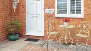 A brick building with a white door, a window with blinds, and a sign reading "Heysham Retreat". There are two metal chairs and a table with a flower pot.