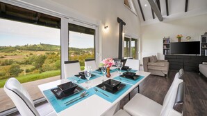 Een eetgedeelte met een tafel voor vier personen, grote ramen met uitzicht op een groen landschap, een bank en een televisie in de woonkamer.