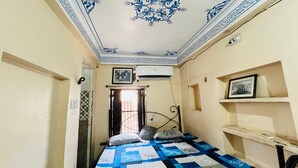 A narrow room with a bed, a ceiling with blue floral patterns, and a wall-mounted air conditioner. - Hotel Heritage haveli (Udaipur)