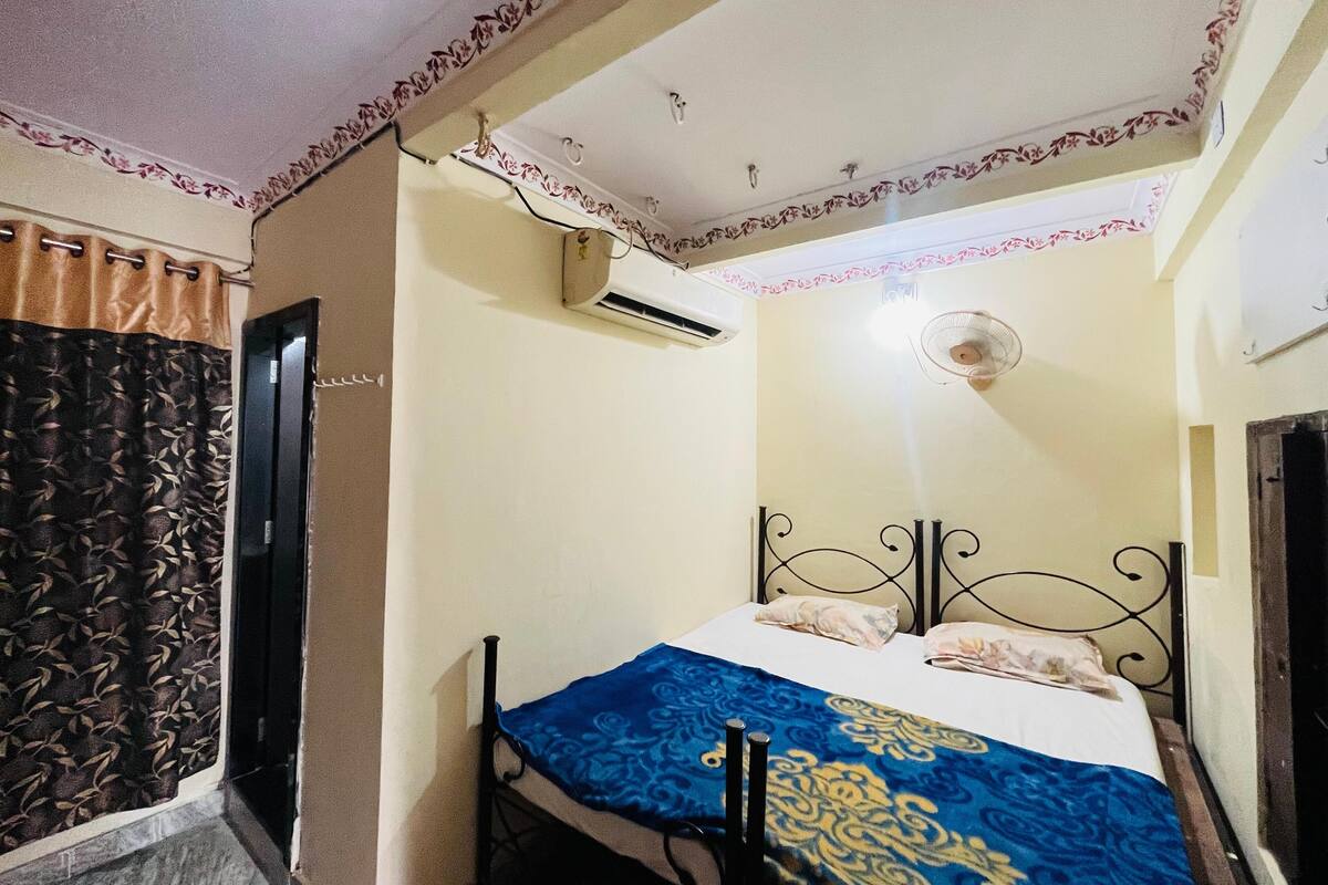 A small, single-bed room with a patterned bedspread, an air conditioning unit, and a ceiling fan.