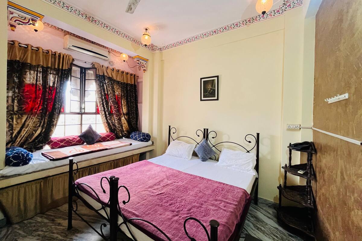 A bedroom with a bed, window with curtains, wall-mounted air conditioner, and a small shelf.