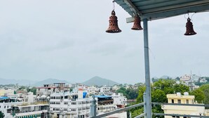 A balcony with hanging bells and a view of a cityscape with buildings and mountains in the background. - Hotel Heritage haveli (Udaipur)