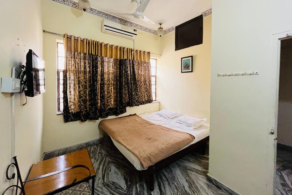 A small, compact room with a bed, a desk, and a chair. There is a window with curtains, an air conditioning unit, and a ceiling fan.