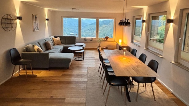 Een ruime woonkamer met een grote houten eettafel, een grijze modulaire bank en uitzicht op een bergachtig landschap door grote ramen.