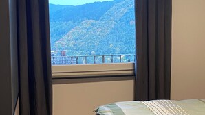 Een slaapkamer met een bed, een raam met gordijnen en uitzicht op een bergachtig landschap.