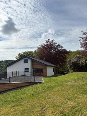 A modern two-story house with a sloped roof, a fenced front yard, and a view of a forested hillside. - Vacation home Natur.Zeit on the Eifelsteig - active - relax - enjoy (Simmerath)