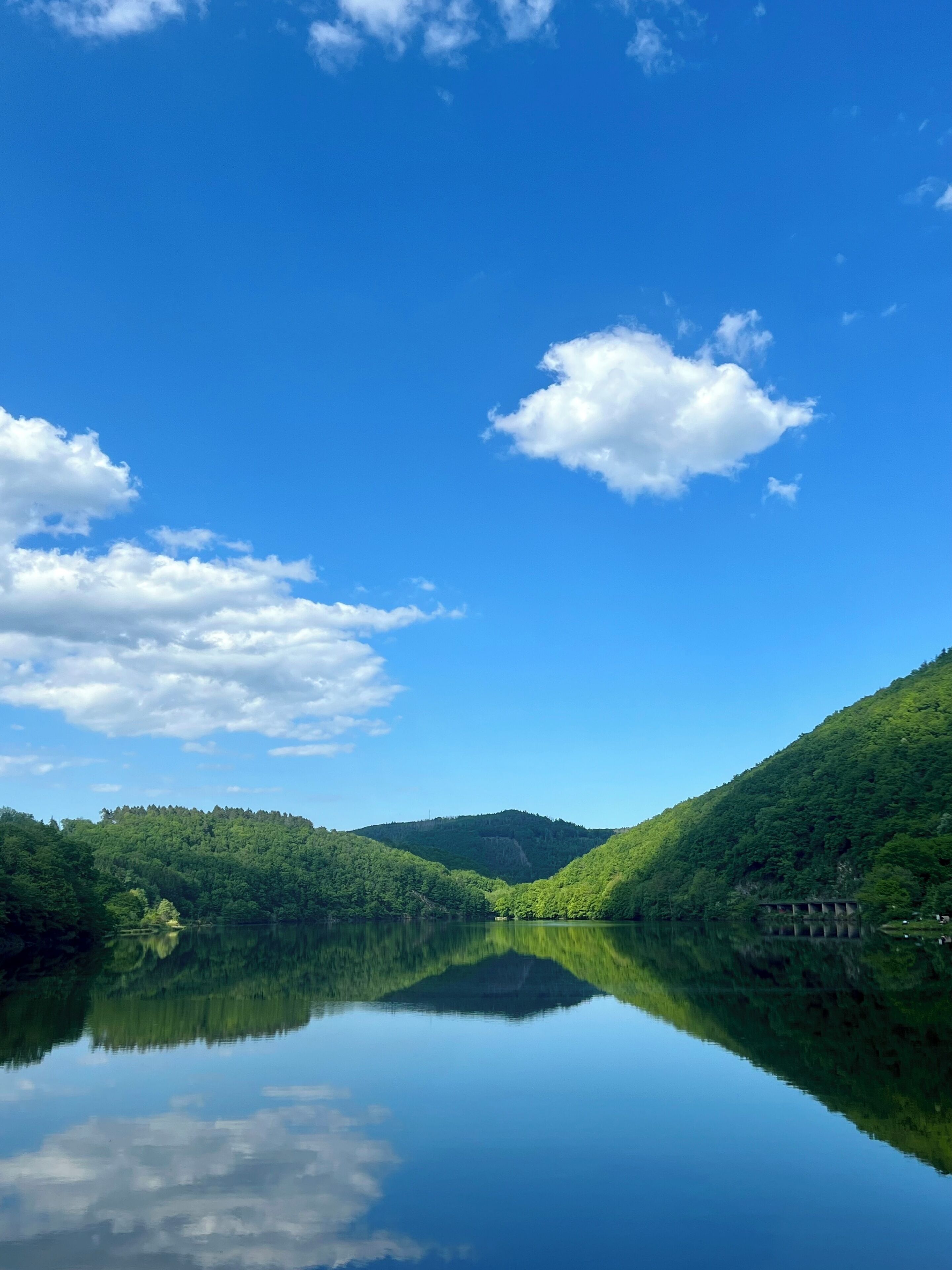 Un lac paisible aux eaux limpides, dont le reflet se mire dans les collines verdoyantes qui l’entourent, sous un ciel bleu.