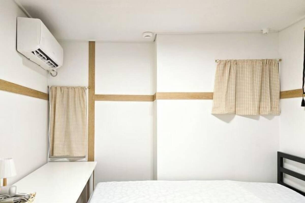 Ein Schlafzimmer mit Bett, Schreibtisch, einem Fenster mit Vorhängen und einer Klimaanlage.