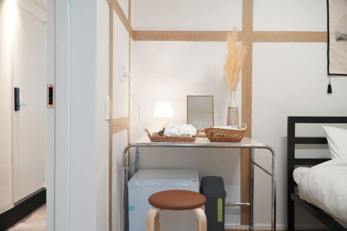 Ein kleines Hotelzimmer mit Bett, einem kleinen Tisch mit Lampe, einem Wäschekorb mit Handtüchern und einem Mülleimer.