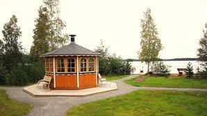 Ein hölzernes Pavillon mit Ziegeldach, umgeben von einem Kiesweg und einer Rasenfläche, mit Blick auf ein Gewässer.