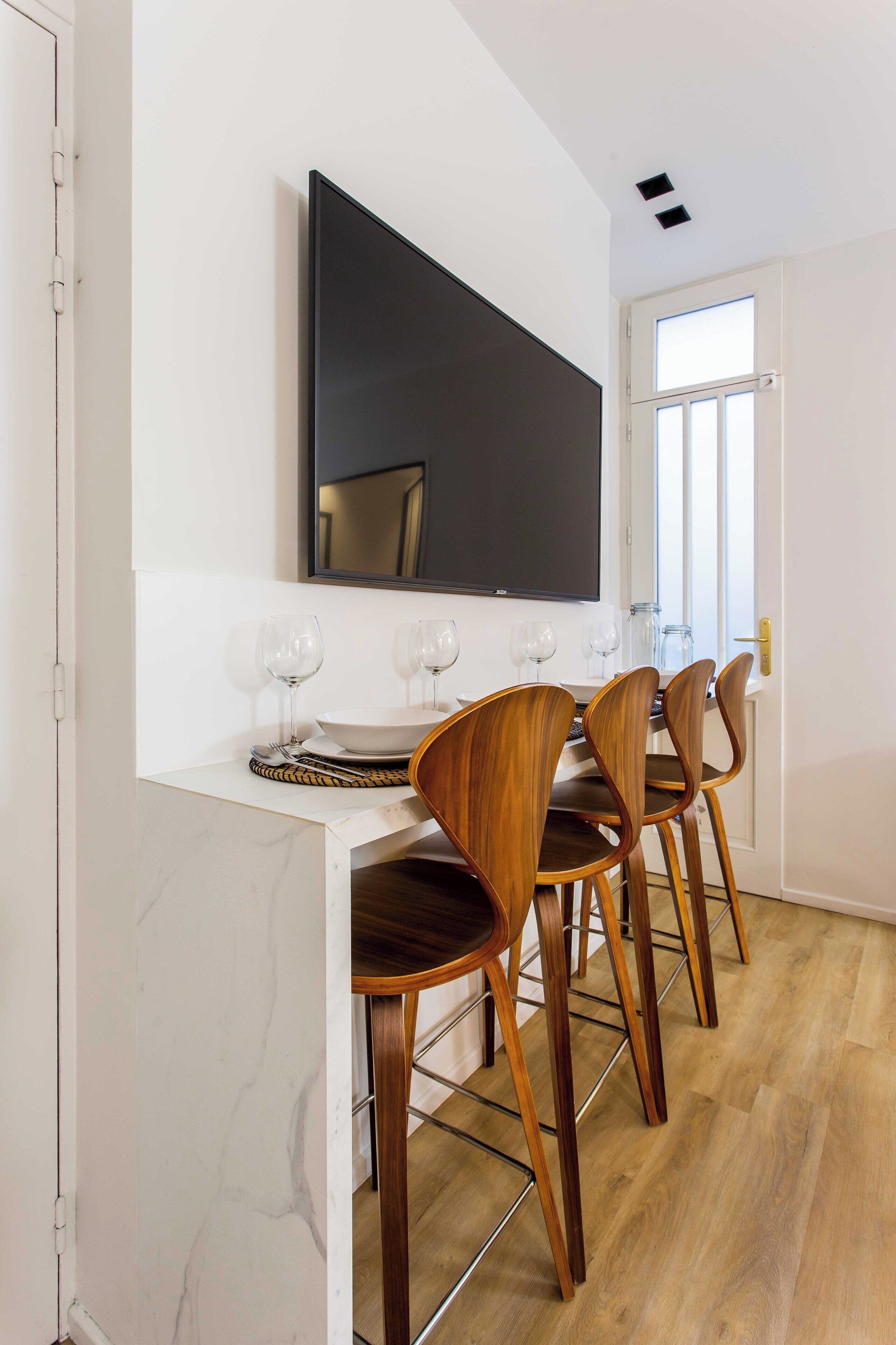Un comedor con encimera de mármol blanco, cuatro sillas de madera y un televisor de pantalla plana grande montado en la pared.