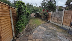A paved backyard with a wooden fence, a small garden area, and a visible house in the background.