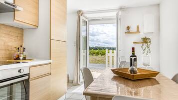 Een moderne keuken met houten kastjes, een eettafel met een dienblad vol wijnglazen en flessen, en uitzicht op een wijngaard door een open deur.