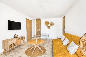 A living room with a TV, wooden furniture, a round table, a sofa with patterned cushions, and wall decor.