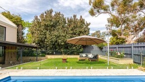 A backyard with a swimming pool, a wooden deck, a lawn, and a large tree.
