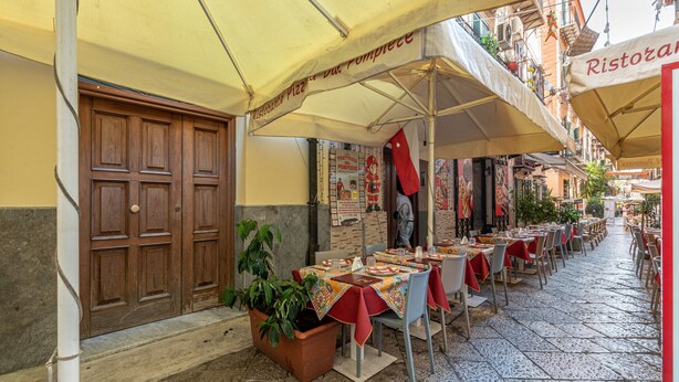 Un área de comedor al aire libre con mesas y sillas bajo sombrillas, una puerta de madera y una planta en maceta.