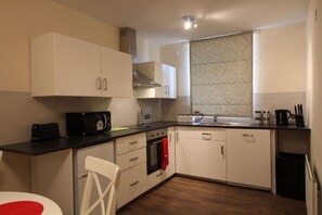 A compact kitchen with white cabinets, a black countertop, a microwave, a stove, and a window with patterned curtains. - Benny's Hideout - City Centre - Parking - 2BR (Belfast)