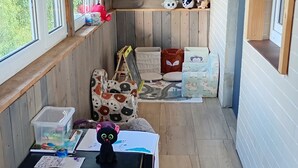 A cozy room with wooden flooring, a black table with a vase and papers, colorful bunting, and various plush toys and cushions.