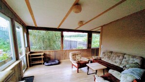 A cozy living room with wooden floors, a sofa, armchairs, and a small table. Large windows offer a view of greenery outside.