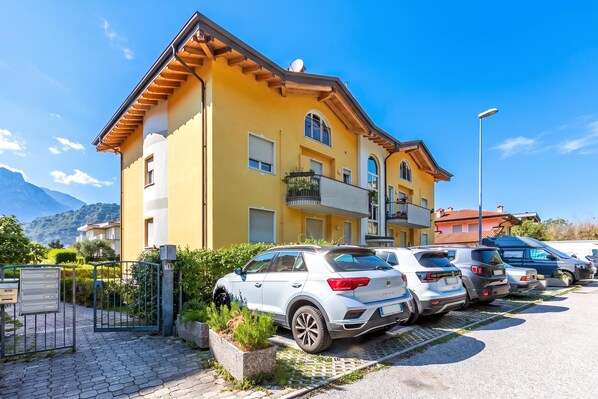 A multi-story building with a yellow facade, parked cars, and a mountainous backdrop. - Torbole On the Moon Apartment Near Beach (Nago-Torbole)