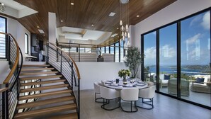 Een ruim interieur met een houten trap, een ronde eettafel gedekt voor zes personen, en een groot raam met uitzicht op zee.