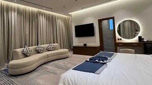 Ein modernes Hotelzimmer mit einer gebogenen Couch, einem großen Bett, einem Flachbildfernseher, einem runden Spiegel und einem Schreibtisch mit Lampe.