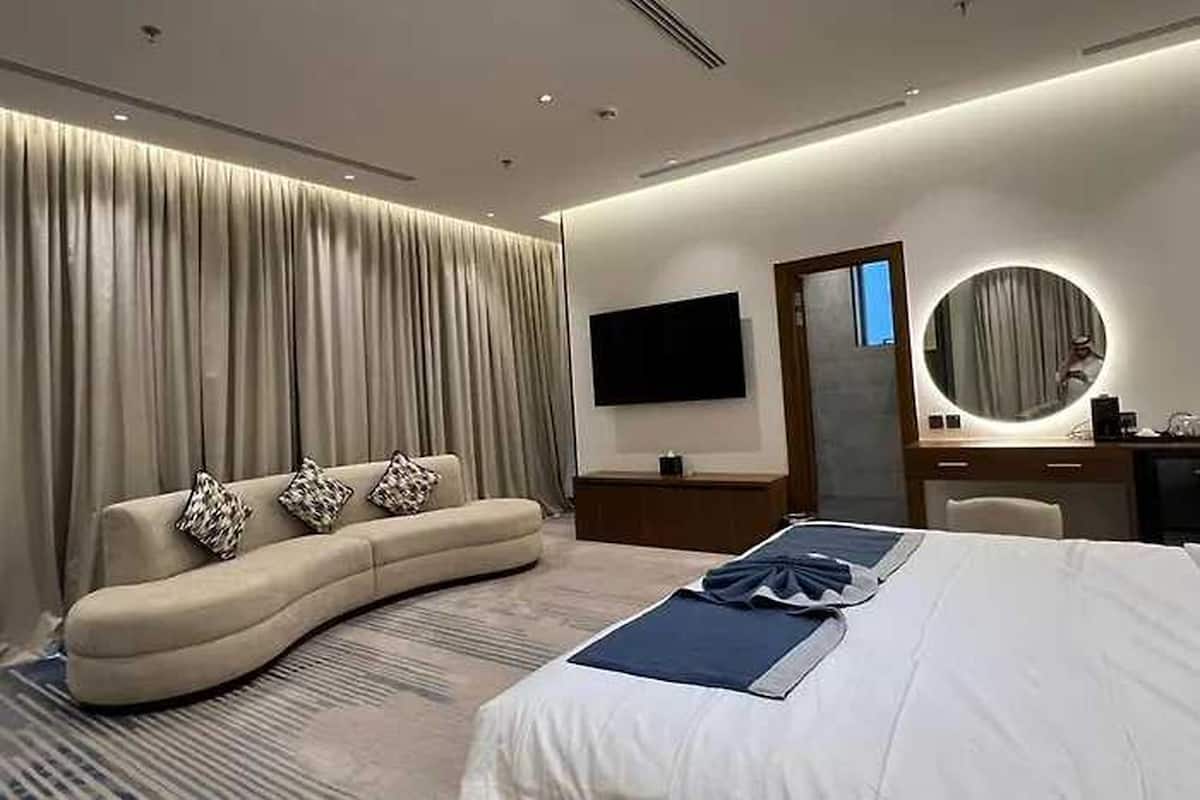 A modern hotel room with a curved sofa, a large bed, a flat-screen TV, a round mirror, and a desk with a lamp.
