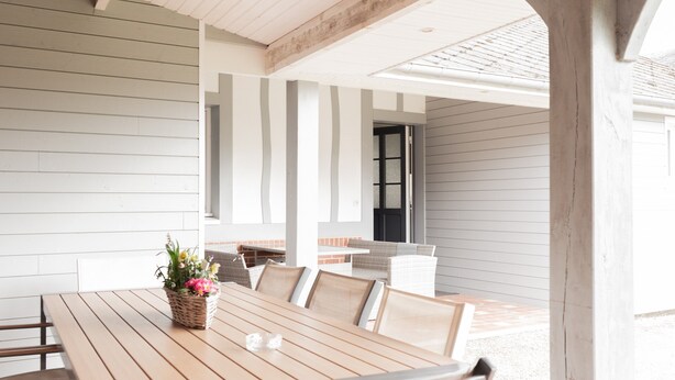 An outdoor dining area with a wooden table and chairs, a basket of flowers, and a view of the house in the background.