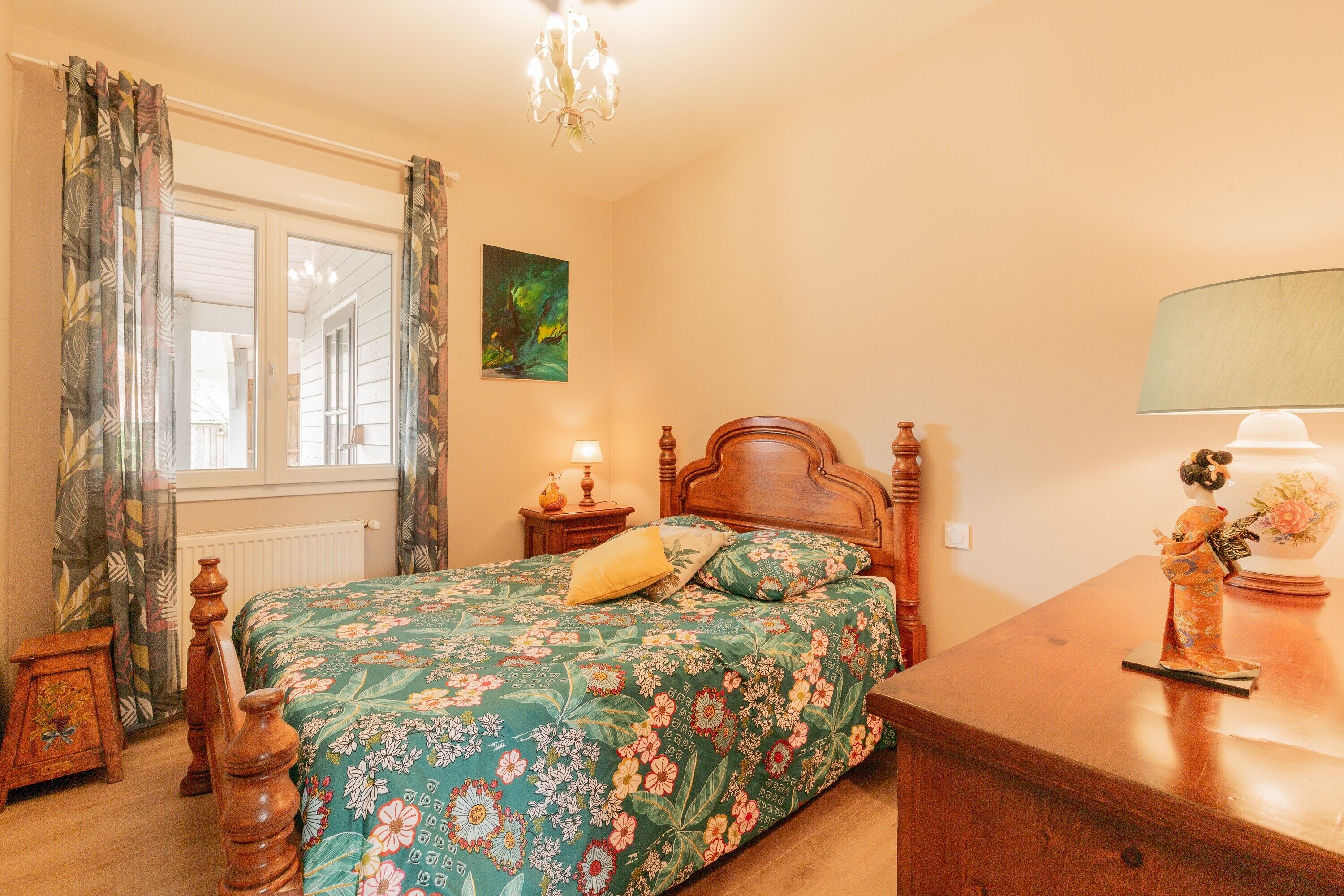 A bedroom with a wooden bed, a nightstand with a lamp, a dresser, and a window with curtains.