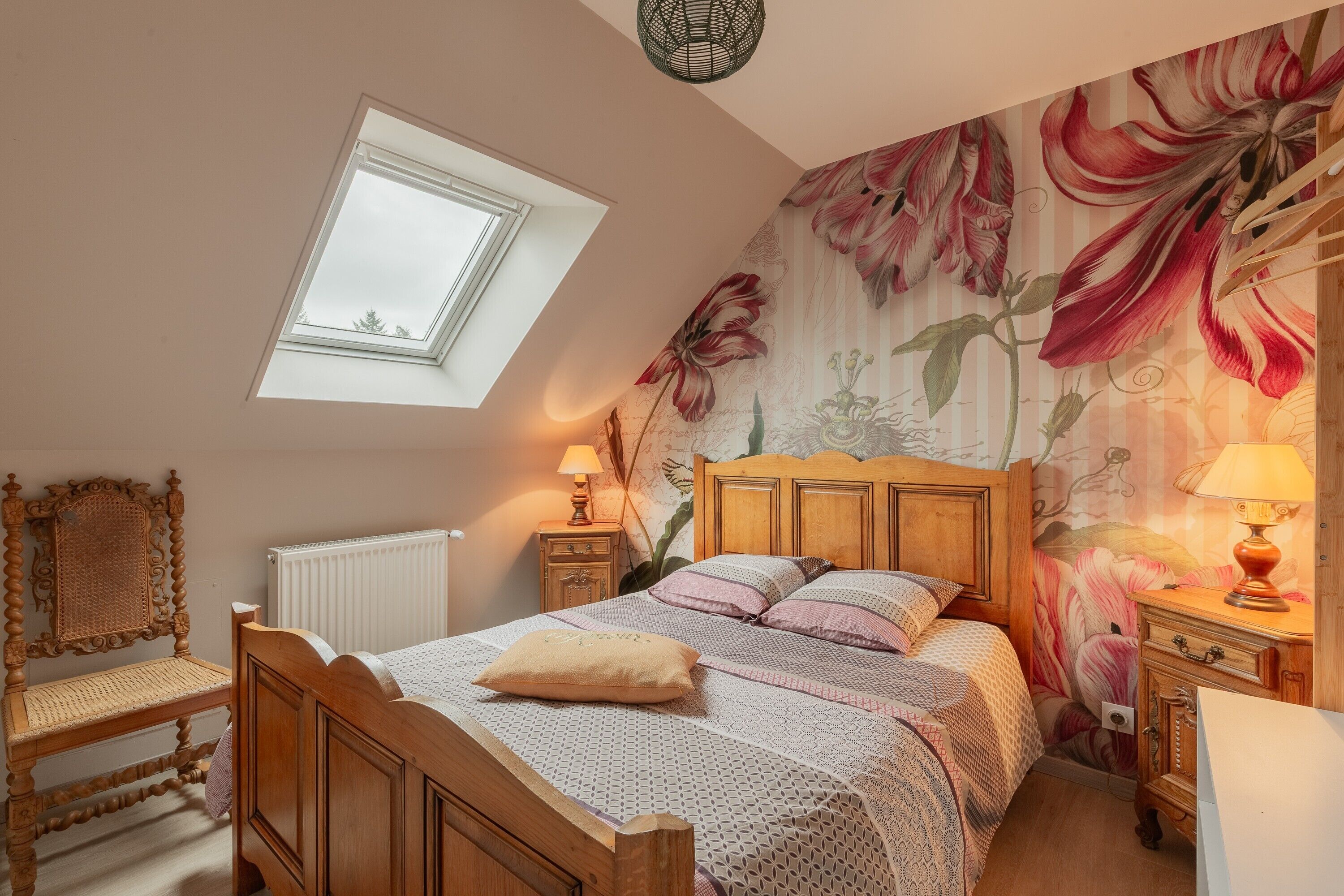 Ein Schlafzimmer mit schrägen Wänden, einem Oberlicht, einem Holzbett, einem Nachttisch, einem Sessel und blumigem Wandpapier.