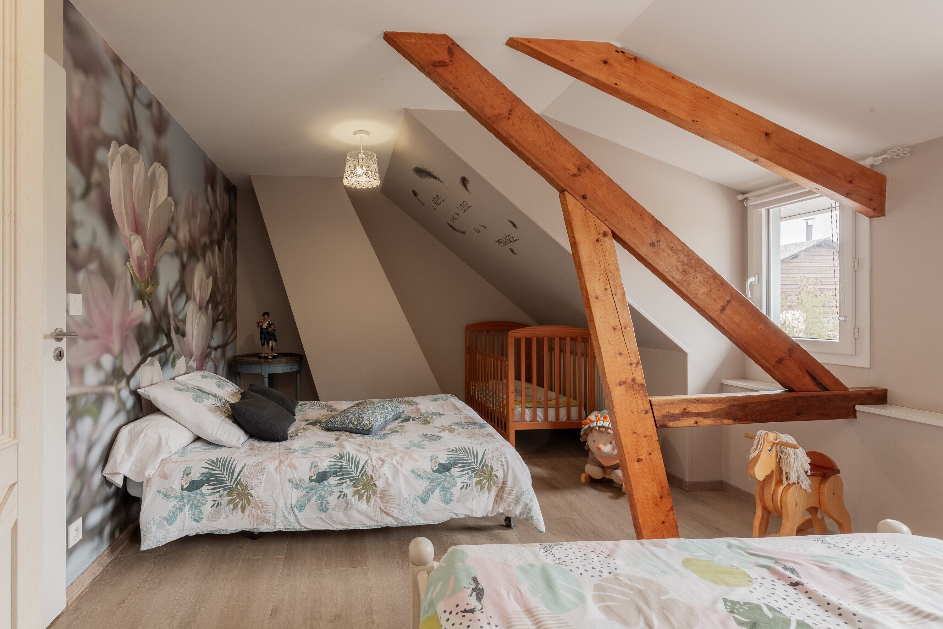 A bedroom with a sloped ceiling, a crib, a rocking horse, and a wall with a floral mural.