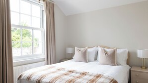 Ein Schlafzimmer mit einem großen Bett, zwei Nachttischen mit Lampen, einem Fenster mit Vorhängen und Blick ins Grüne.