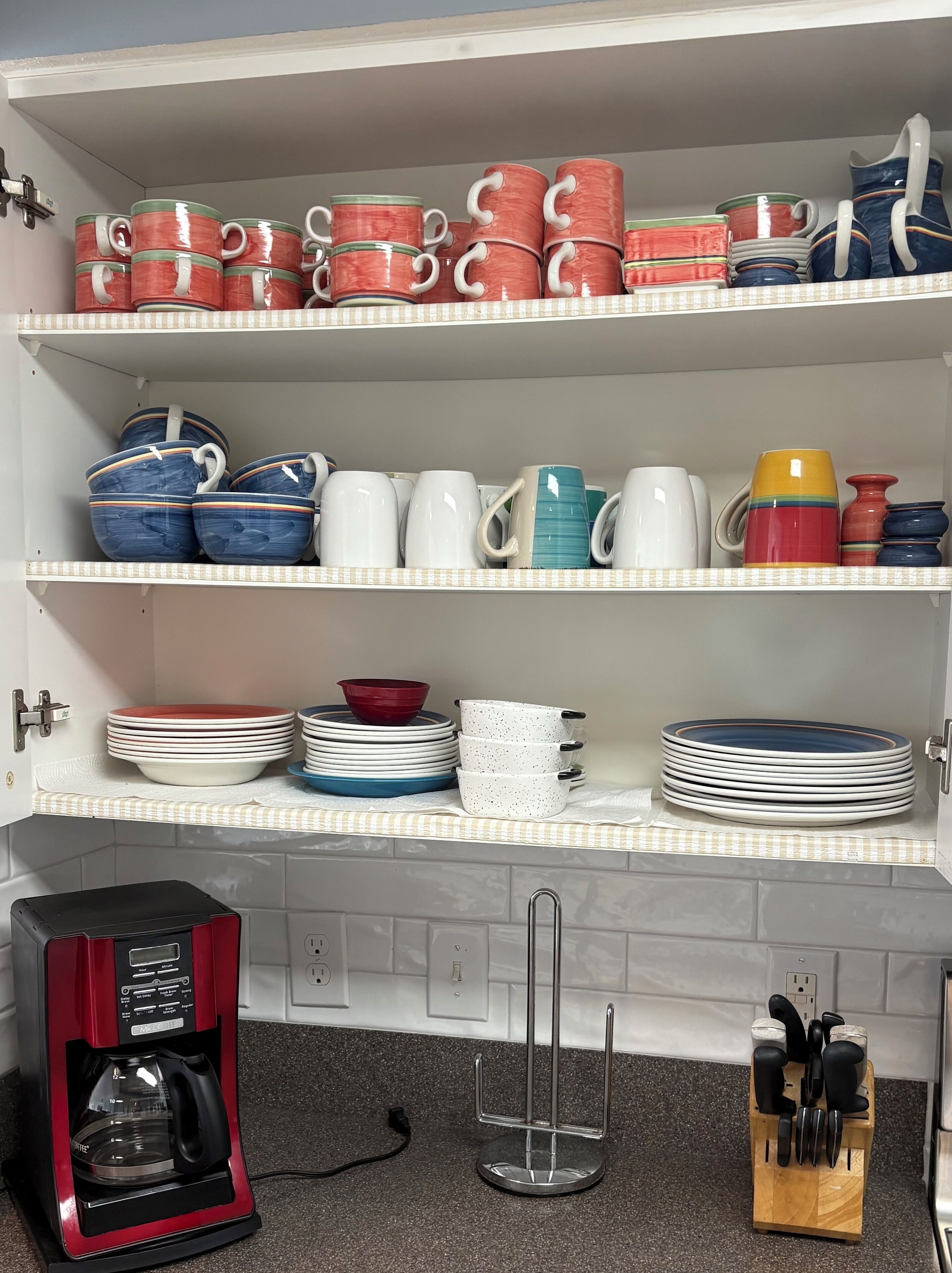Una cocina con estantes que sostienen varias tazas y cuencos, una cafetera y un bloque de cuchillos.