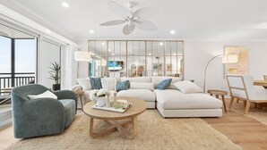 Ein modernes Wohnzimmer mit einem Ecksofa, einem grauen Sessel und einem Couchtisch. Ein Deckenventilator, ein großes Fenster und ein Blick ins Freie sind ebenfalls vorhanden.
